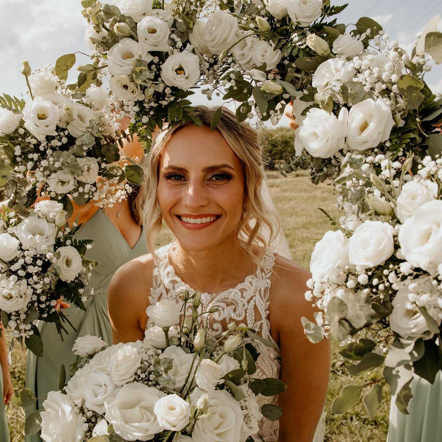 Woman smiling surrounded by white roses and greenery, wearing a lace dress, outdoors.